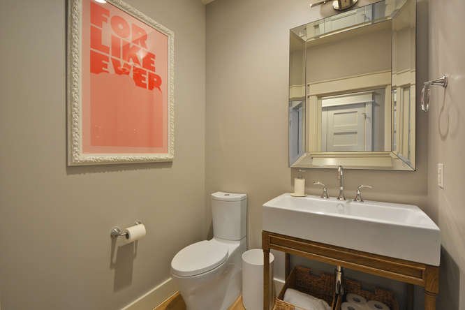 Bathroom with beige walls, framed pink and white artwork, white toilet, oval mirror, small sink with wooden stand, towel ring, and toilet paper holder.