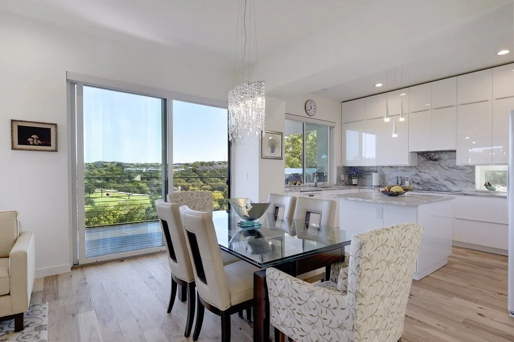 Modern dining area with a glass-top table surrounded by cream upholstered chairs, overlooking a balcony with a scenic view of trees and greenery, and an open white kitchen with marble countertops and hanging pendant lights.