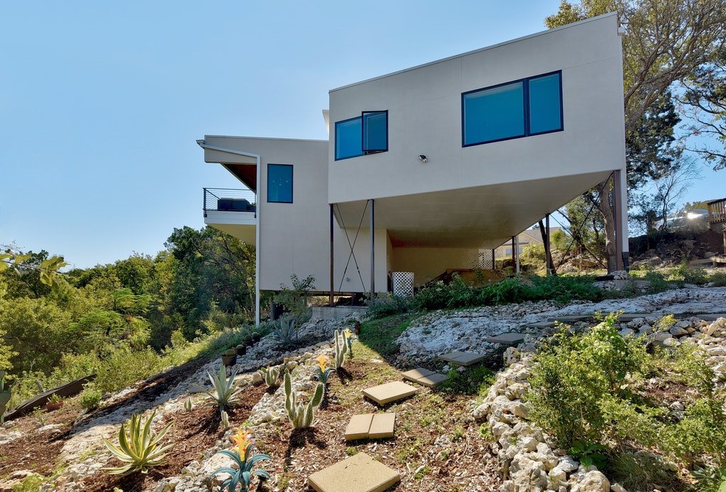 Modern white house built on a rocky hillside with a large overhang and multiple windows, surrounded by desert plants and sparse trees.