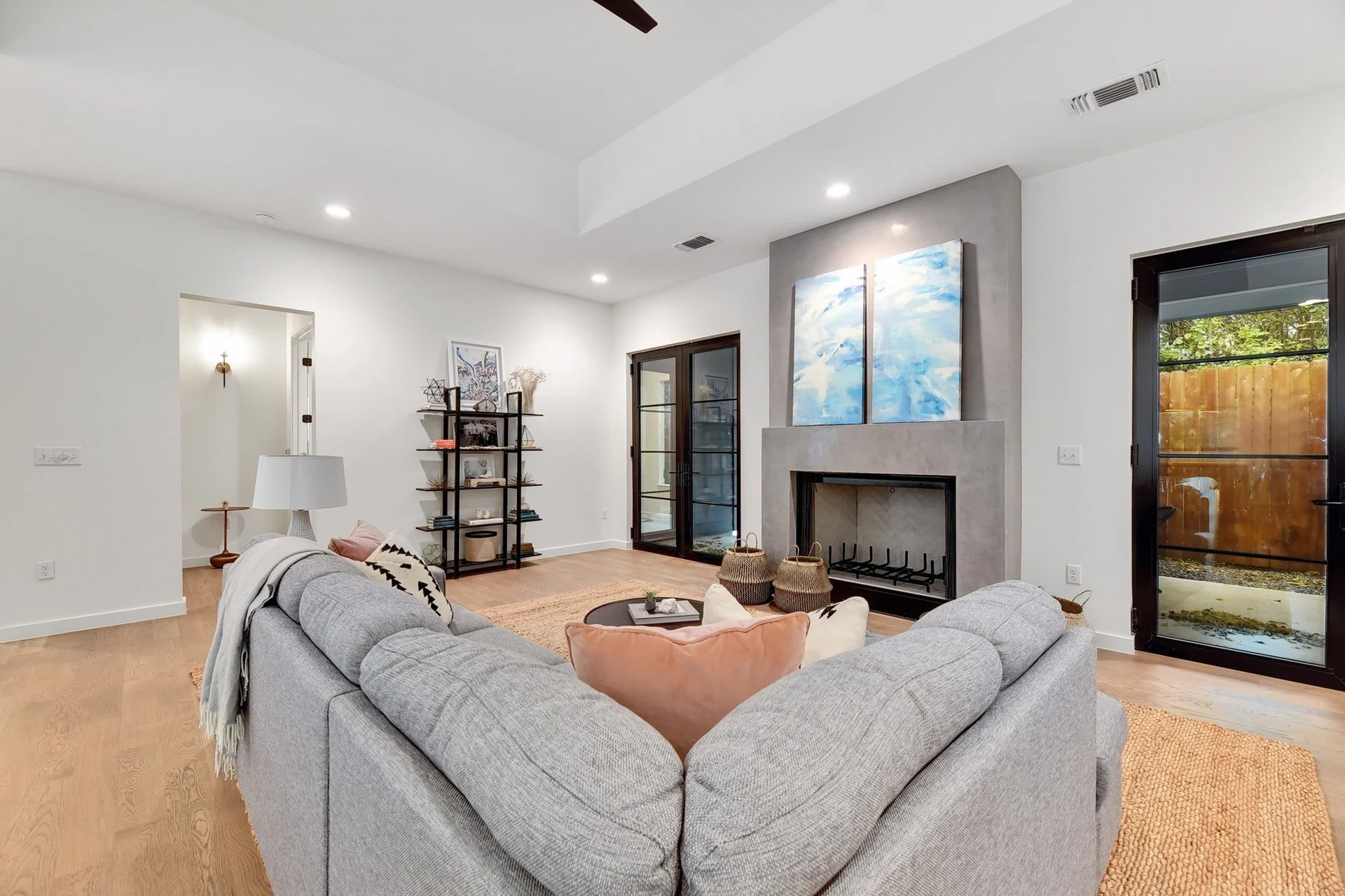 Living room with a gray sectional sofa, a fireplace with abstract blue and white art above, a black sliding glass door, and wooden floors.