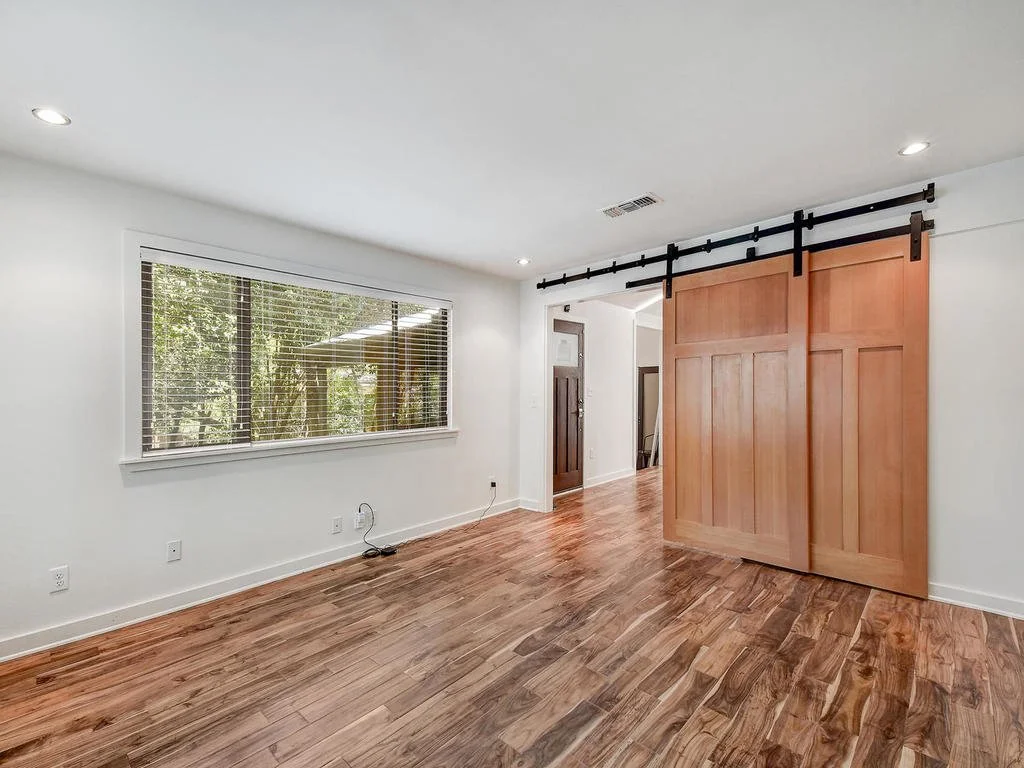 Empty room with hardwood floors, white walls, a large window with blinds, and a wooden sliding barn door.