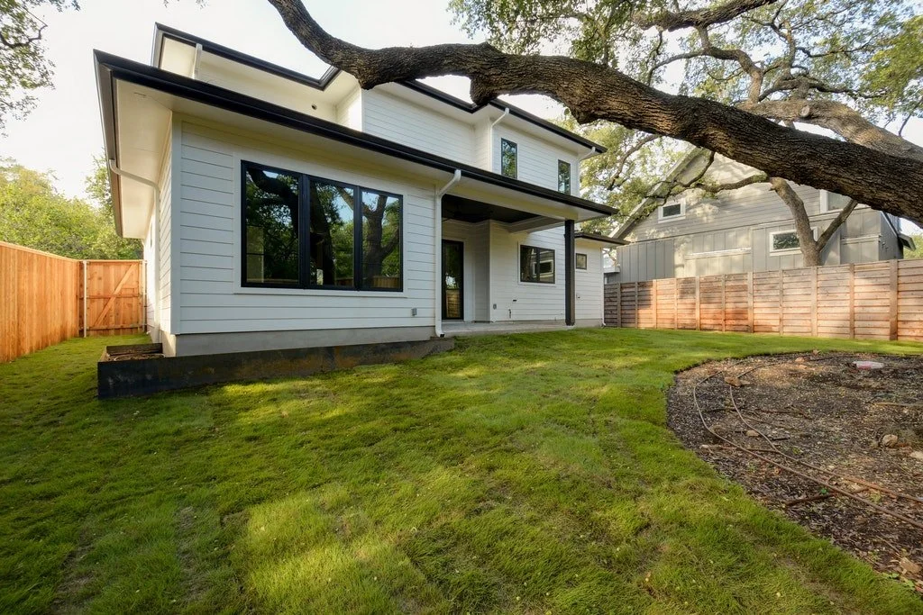 White two-story house with modern design, black window frames, and a large tree in the front yard, on a sunny day.