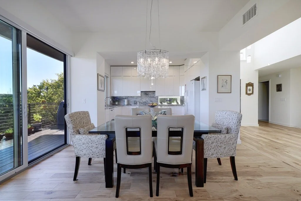 Modern open-concept dining area with a rectangular glass table, central chandelier, and six chairs, adjacent to a kitchen with white cabinets and a marble backsplash, and a sliding glass door leading to a balcony with outdoor seating.