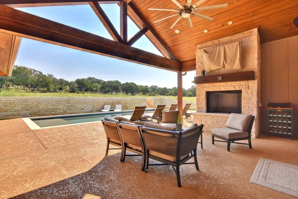 Indoor outdoor living space with fireplace, ceiling fan, and patio furniture leading to a backyard with a pool and trees.