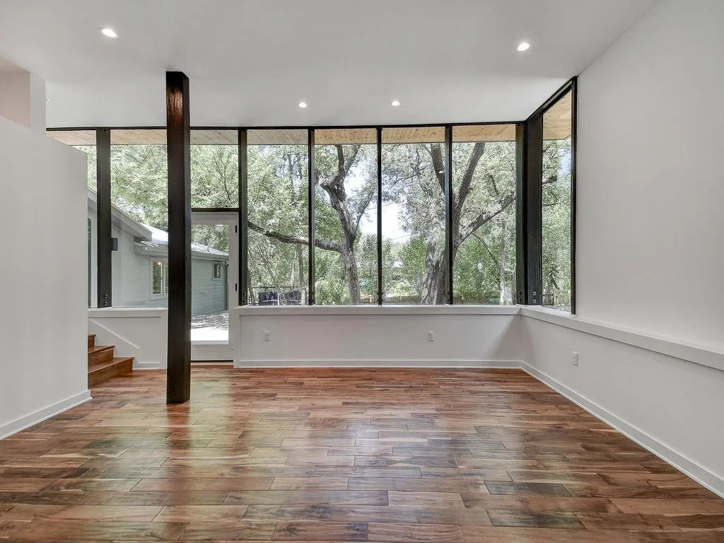 Empty room with large windows showing trees outside, hardwood floors, white walls, and recessed ceiling lights.