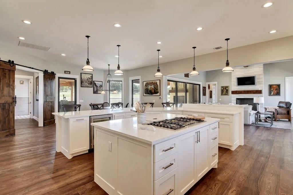 Open-concept kitchen with white island and pendant lighting, adjacent dining area and living room with fireplace, wooden floors, and sliding glass doors.
