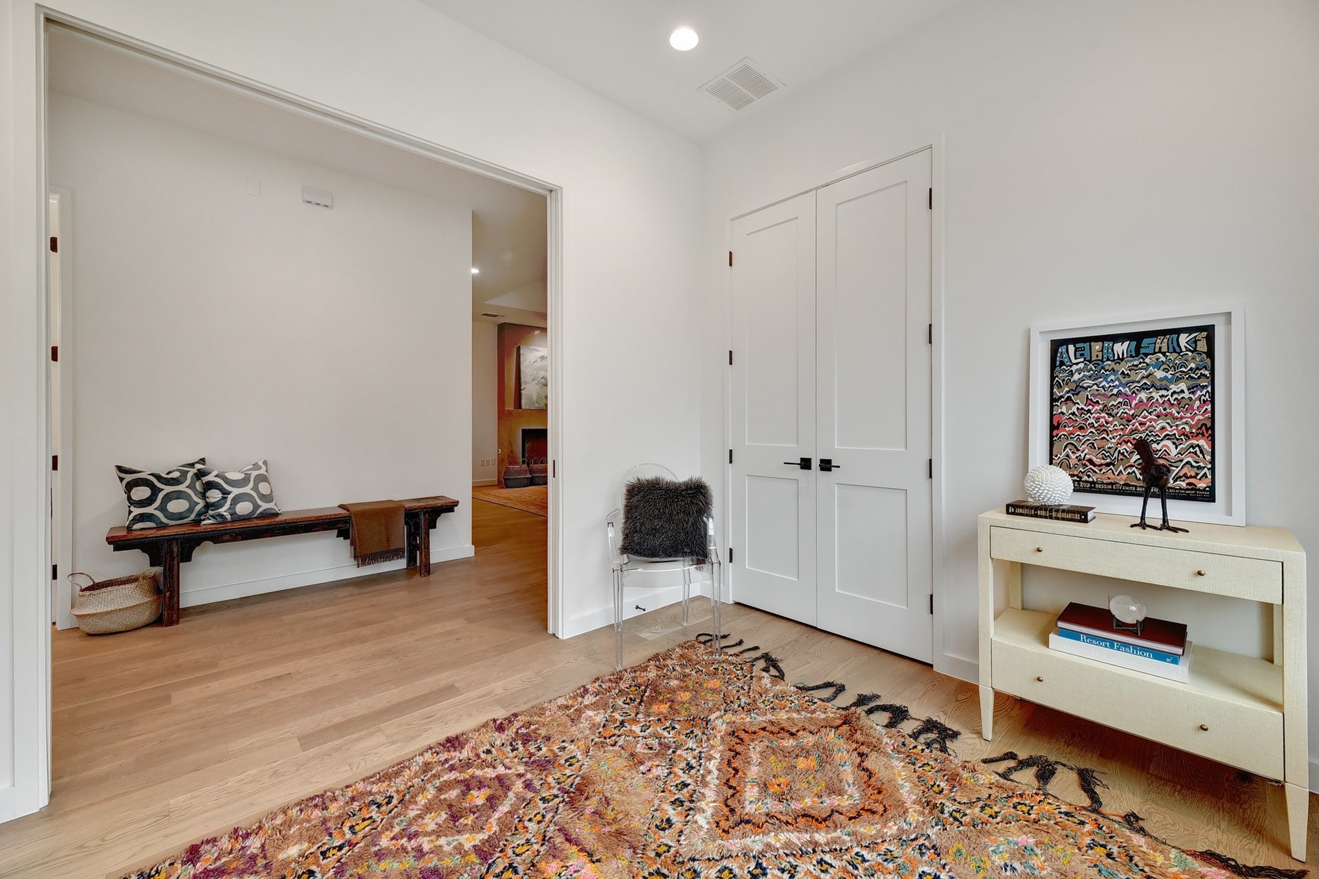 Living room with white walls and wooden floor, featuring a colorful patterned rug, a white shelf with books, colorful wall art, and a bench with pillows.