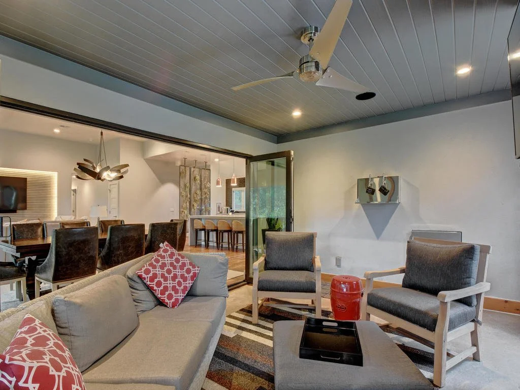 Living room with gray sofa and two armchairs, wall-mounted shelves, red ceramic side table, and a chandelier dining area in the background.