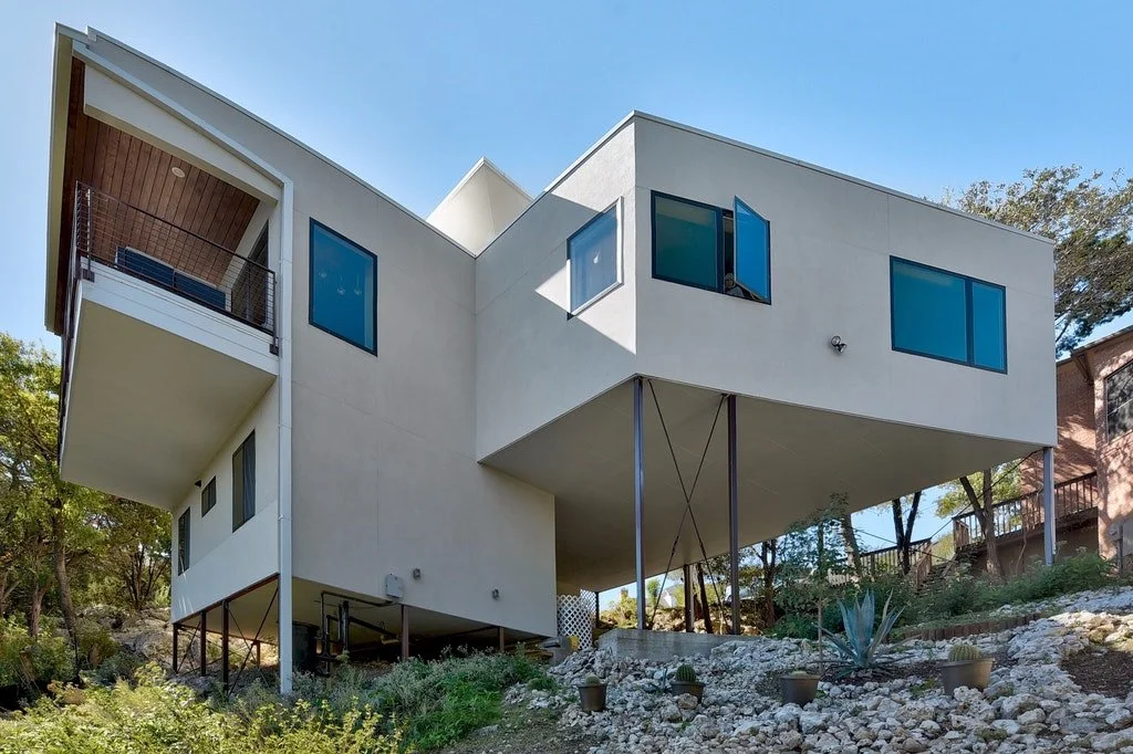 Modern house with a white exterior, elevated on stilts, with large blue-tinted windows and a small balcony, set against a clear blue sky and surrounded by sparse trees and rocky landscaping.