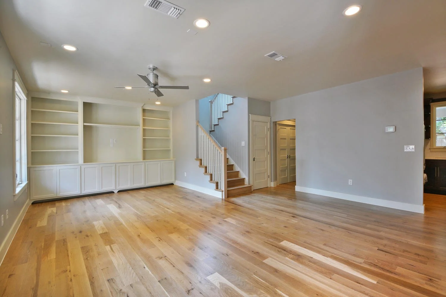 Empty living room with hardwood floors, white walls, built-in white shelves, and a staircase with white railing leading upstairs.