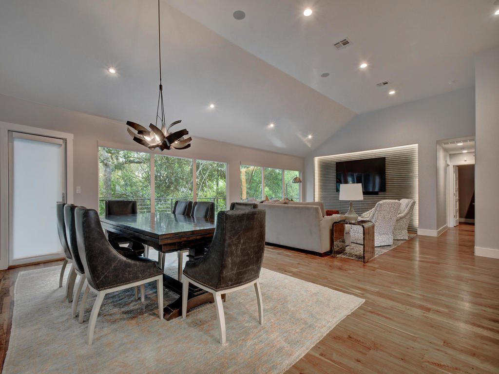 Open-concept living and dining room with large windows, hardwood floors, modern chandelier, black dining table with six chairs, white and patterned sofas, wall-mounted TV, and minimal decor.