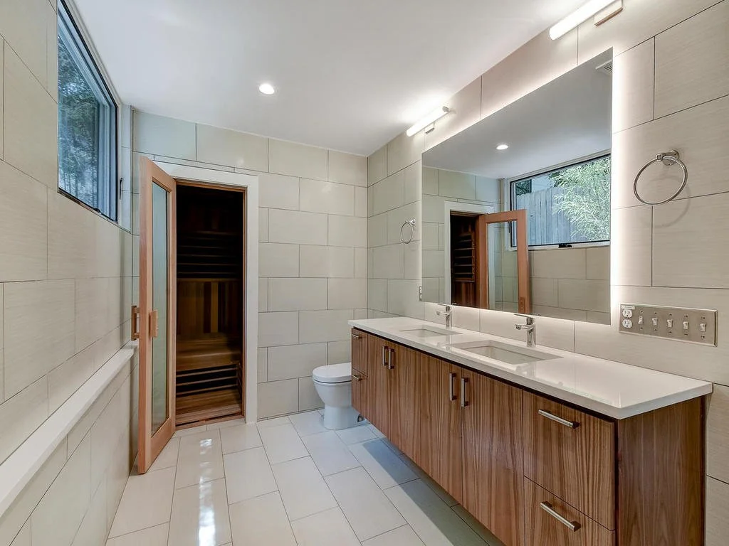 Modern bathroom with dual sinks, large mirror, wooden cabinets, toilet, window, and a sauna.