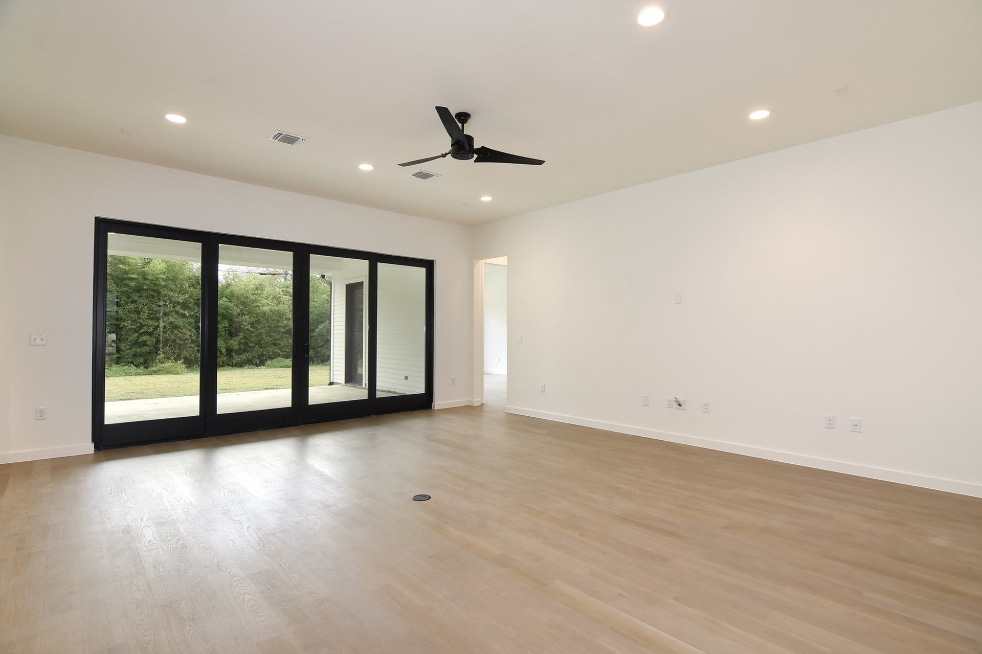 Empty living room with light wood flooring, white walls, black sliding glass doors leading to outdoor area, ceiling fan, and recessed lighting.