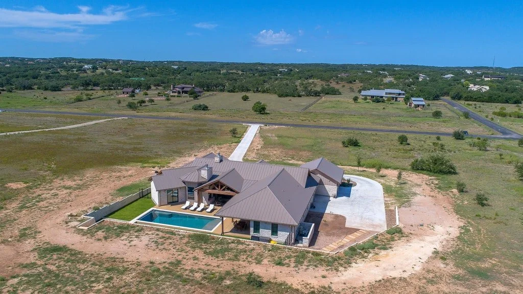 A modern house with a swimming pool in a large, open grassy area. The house has a metal roof and outdoor lounging area, with a concrete driveway leading up to it. Several other houses are visible in the distance under a blue sky.