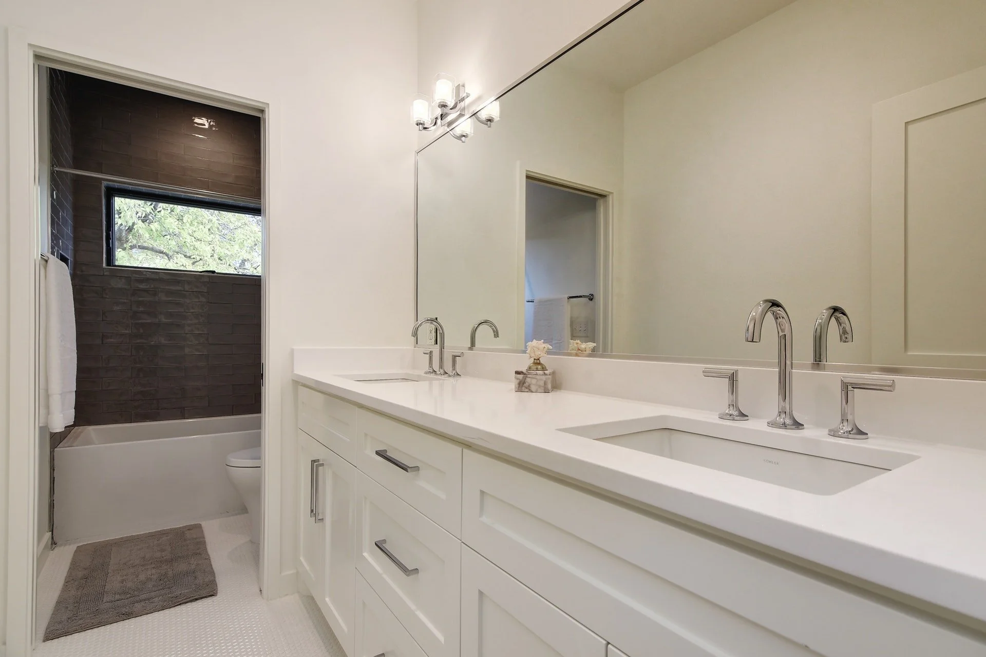 Modern bathroom with white vanity and dual sinks, large mirror, and light fixture. Shower with dark brown tile and window in background.