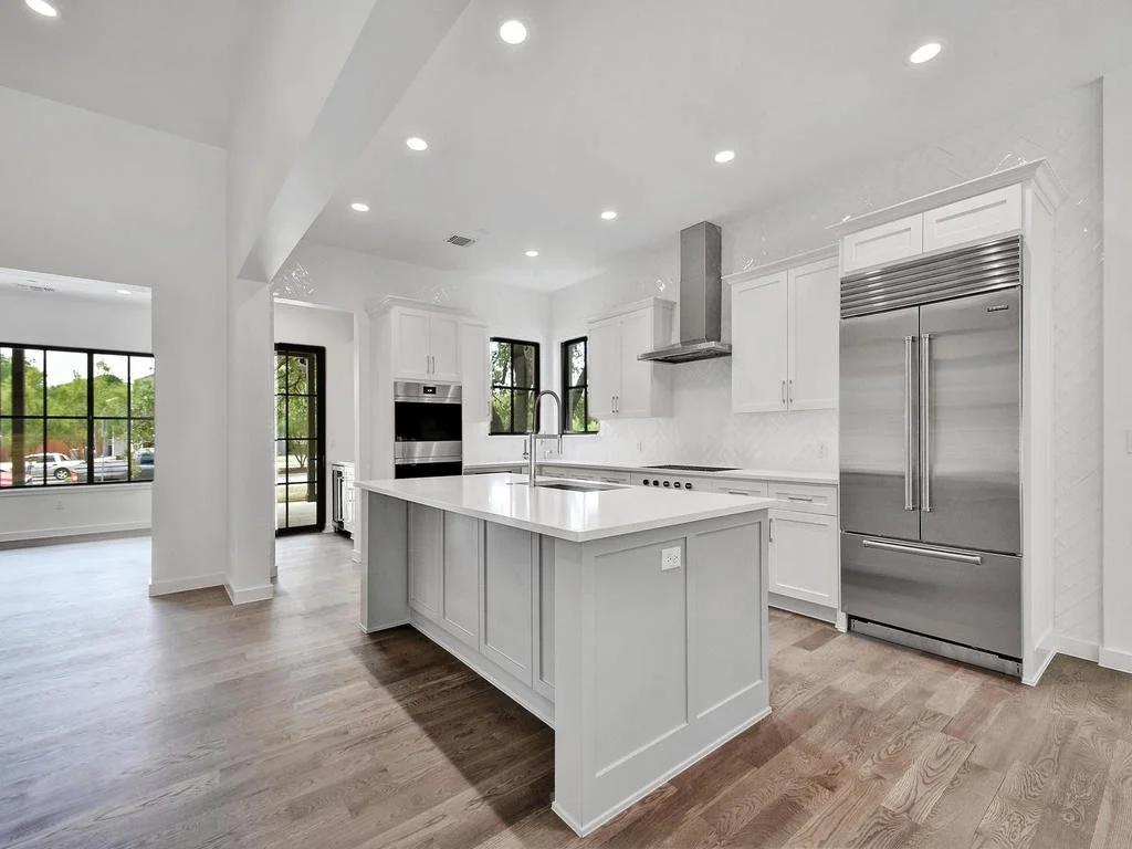 Modern white kitchen with island, stainless steel appliances, hardwood floors, and large windows.