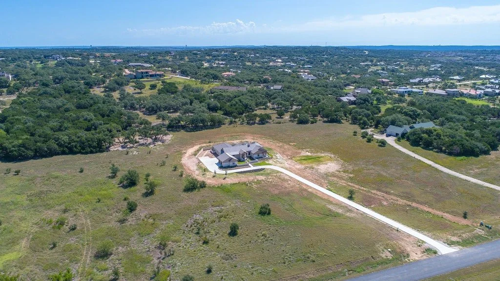 Aerial view of a house under construction surrounded by open fields and trees, with a paved driveway leading to the house in a suburban area.