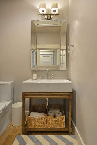 Bathroom with white sink vanity, mirror, wall-mounted light fixture, toilet, woven storage baskets with toilet paper, and a towel ring, all in a narrow space with light-colored walls and wood flooring.