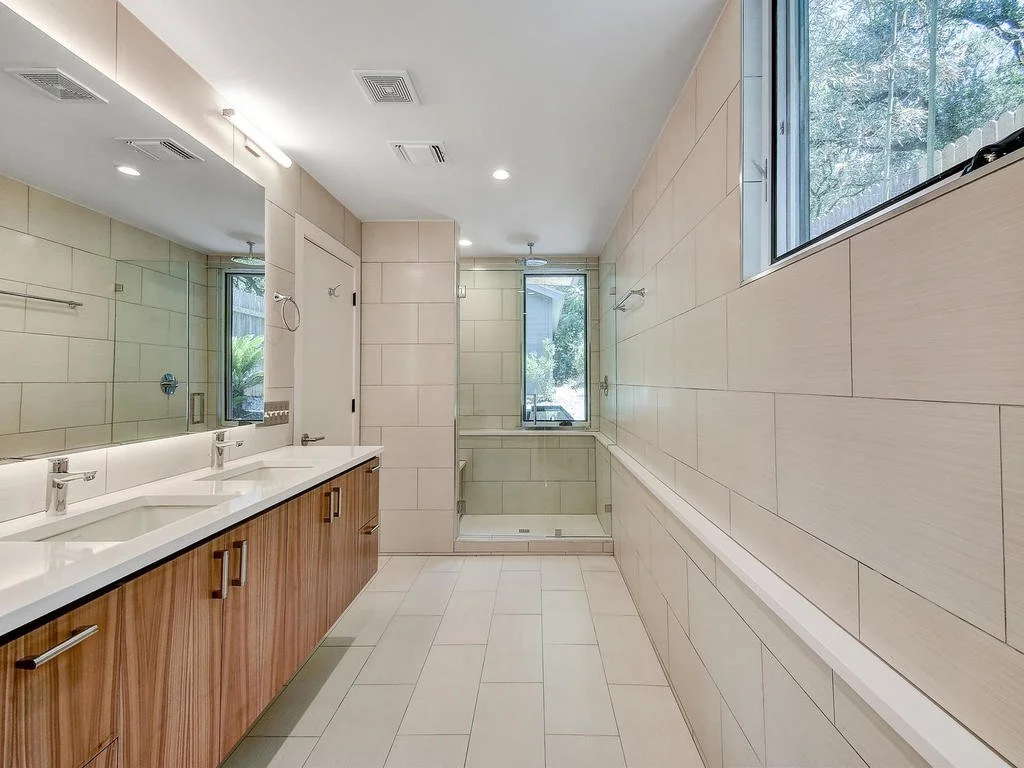 Modern bathroom with dual sinks, large mirror, beige tile walls, and a glass-enclosed shower.
