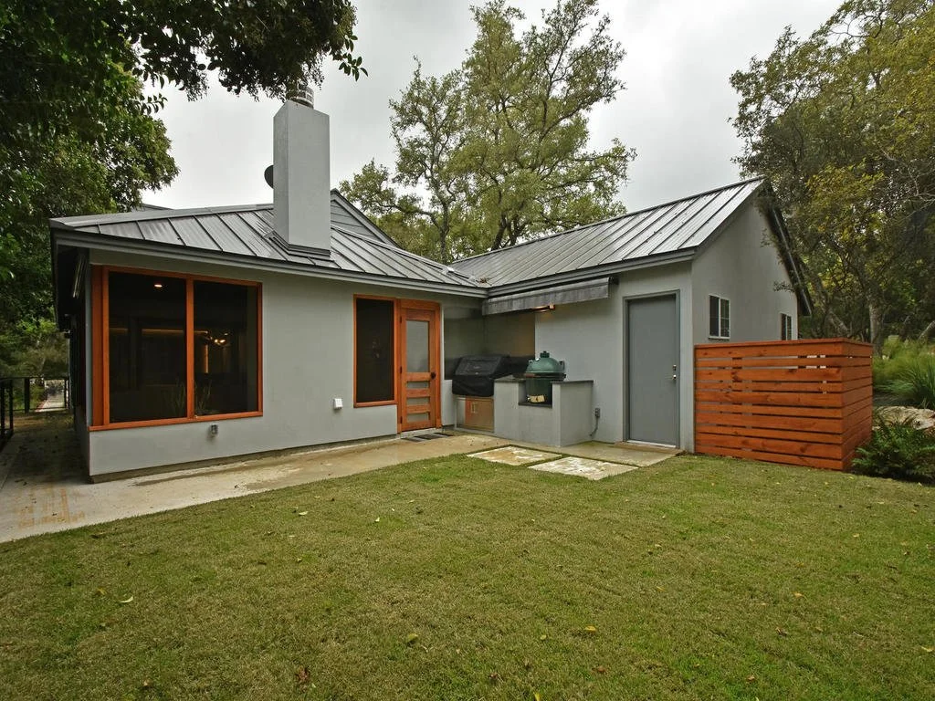 The image shows the back of a modern house with a gray exterior, metal roof, wooden-framed windows, and a fenced backyard with grass and concrete patio.