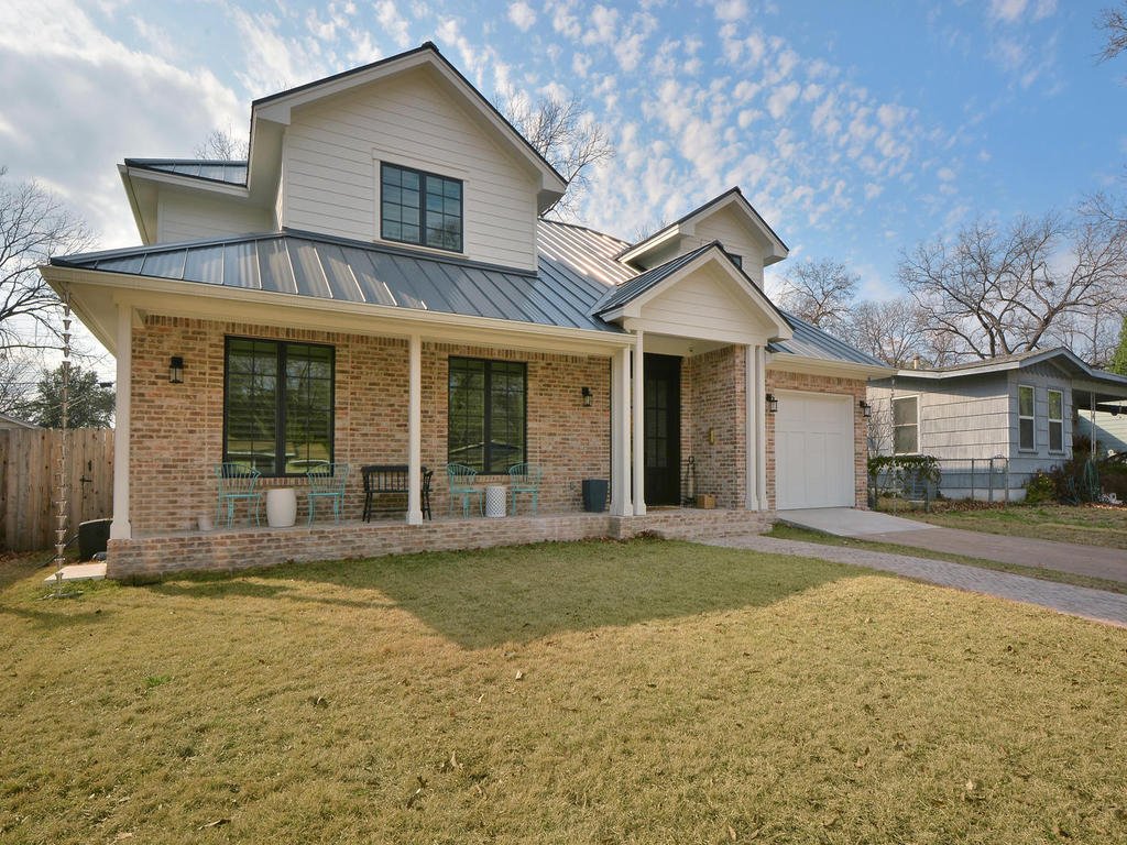 Front view of a two-story house with brick and siding exterior, metal roof, front porch with chairs, and a lawn.