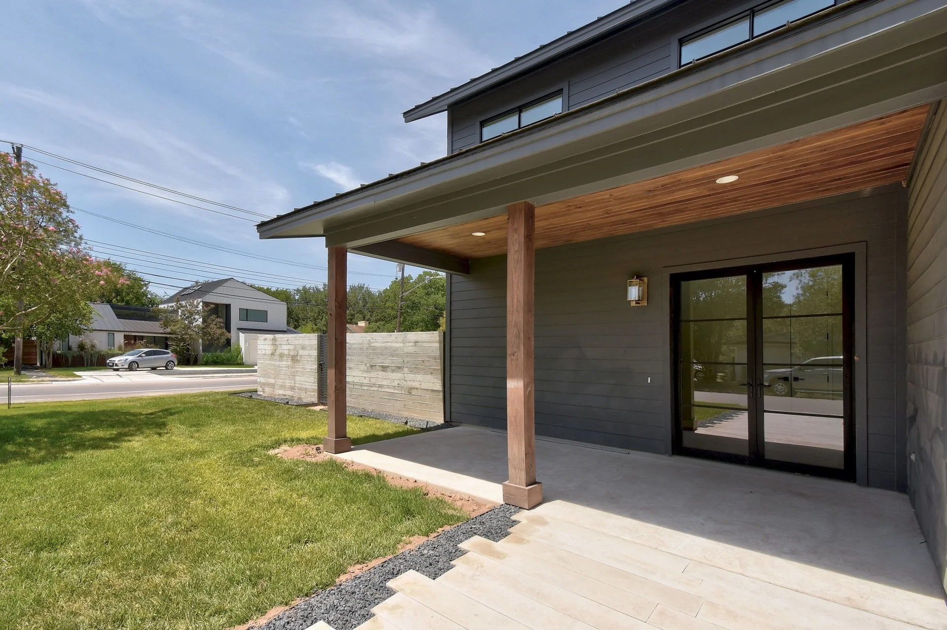 Backyard patio with concrete floor and sliding glass door, covered by a wooden ceiling supported by wooden posts, with a grassy lawn and other modern houses in the background.
