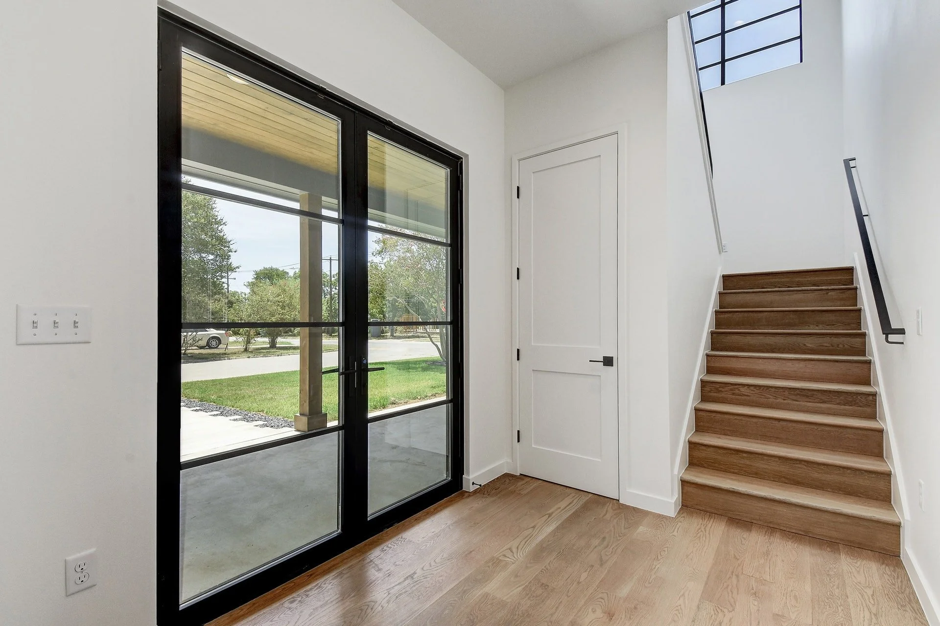 Interior view of a modern house entryway with a black-framed glass door, white walls, a wooden staircase, and a window at the top of the stairs.