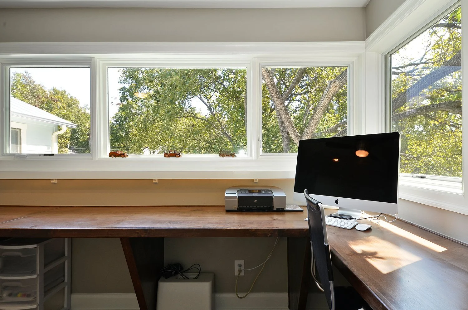 Home office with a wooden desk, computer monitor, printer, and large window with trees outside