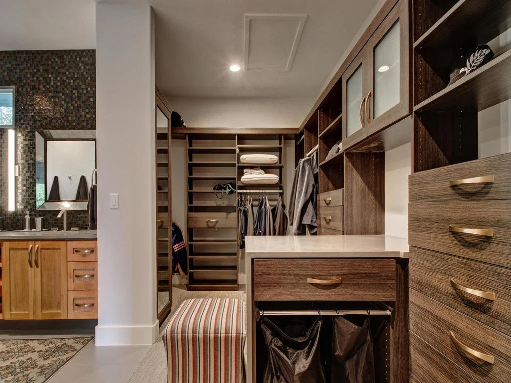 Walk-in closet with wooden shelves, drawers, and hanging space for clothes, adjacent to a bathroom with a mirror and vanity.