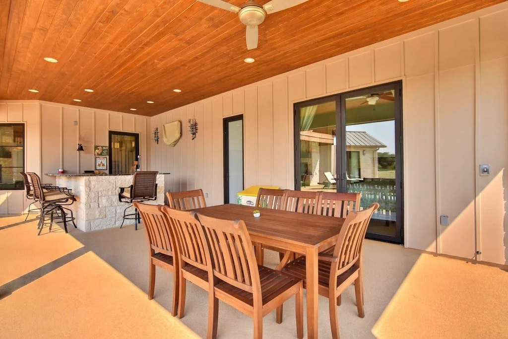 Covered outdoor patio with a wooden dining table and chairs, a bar with stools, and sliding glass doors to the house interior.