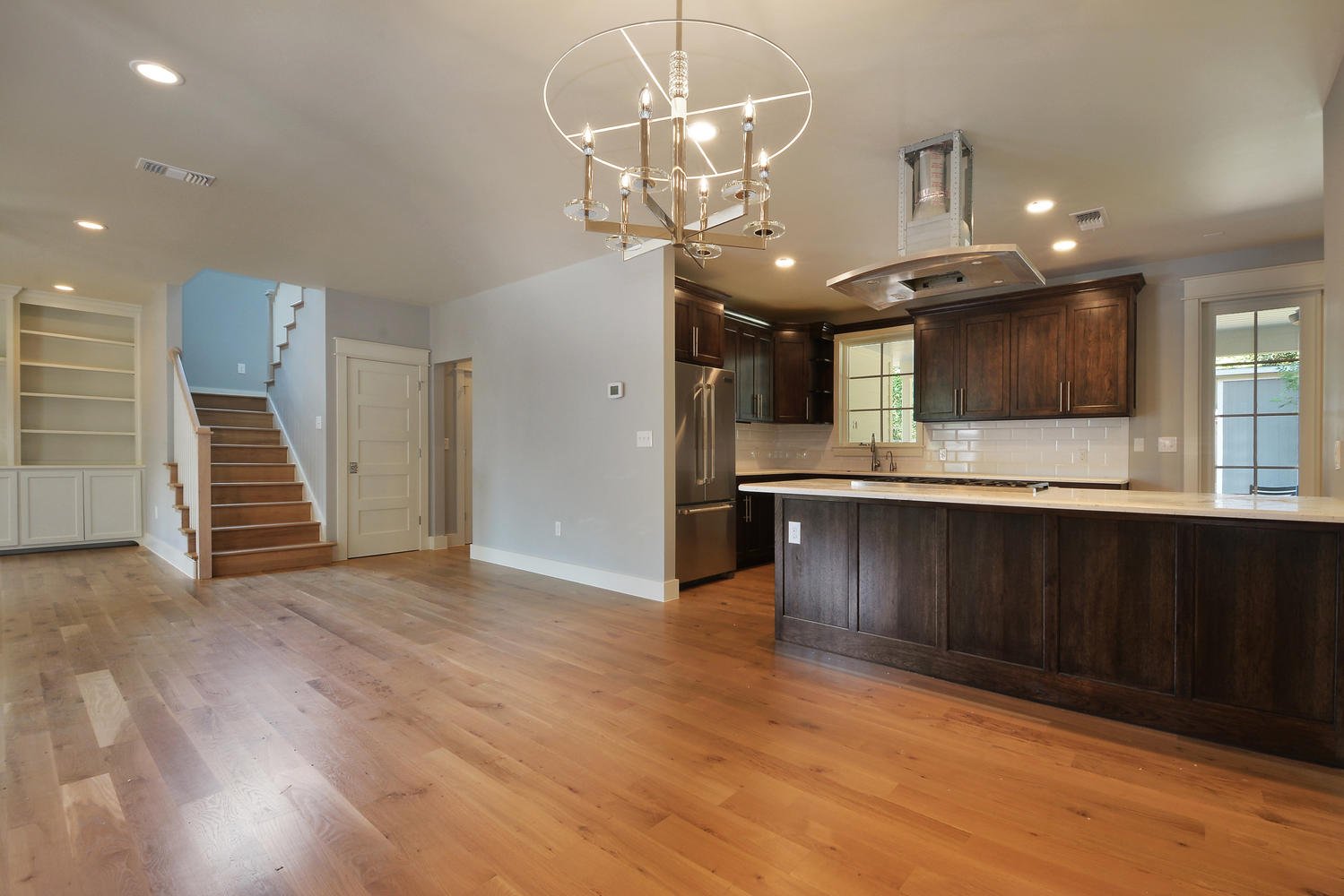 Open-concept living area with hardwood floors, staircase, and kitchen featuring dark wood cabinets, white countertops, stainless steel appliances, and a chandelier overhead.