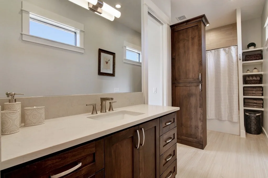 Bathroom vanity with a sink, wooden cabinetry, small framed art on wall, two small high windows, tall wooden storage cabinet, shelves with towels, and a shower with a curtain.