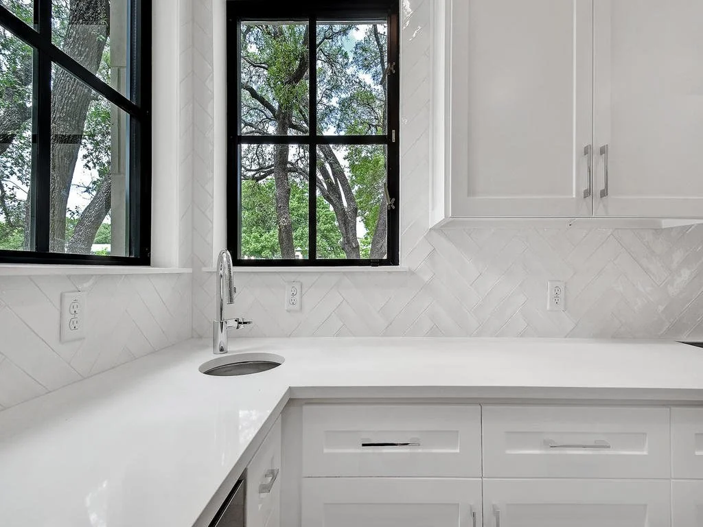 White kitchen countertop with an integrated sink, silver faucet, and white cabinetry below. Two black-framed windows showing green trees outside, with natural light illuminating the space.