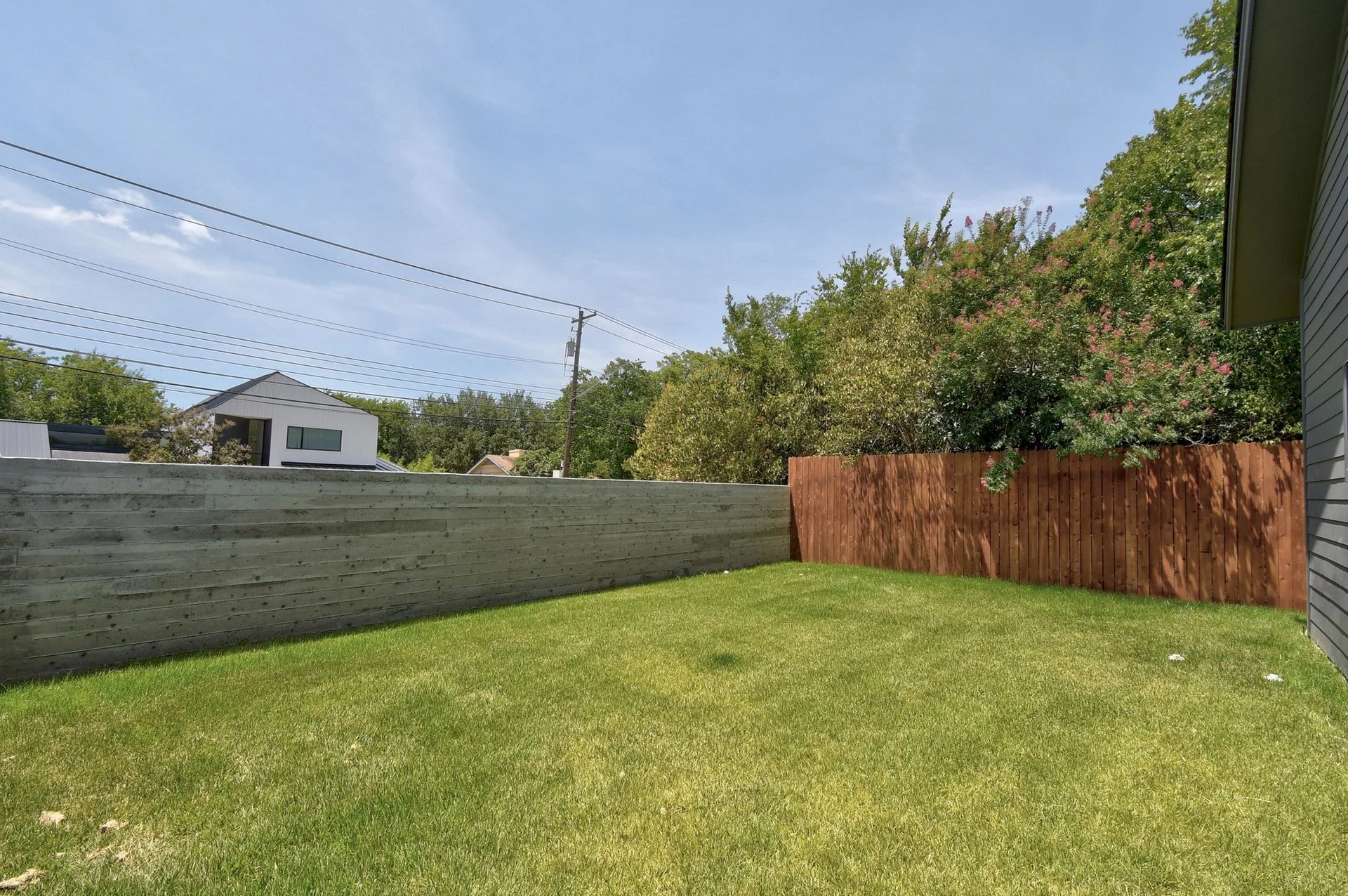 Backyard view with green lawn, wooden fence, trees with pink flowers, house wall on the right, and clear blue sky with power lines overhead.