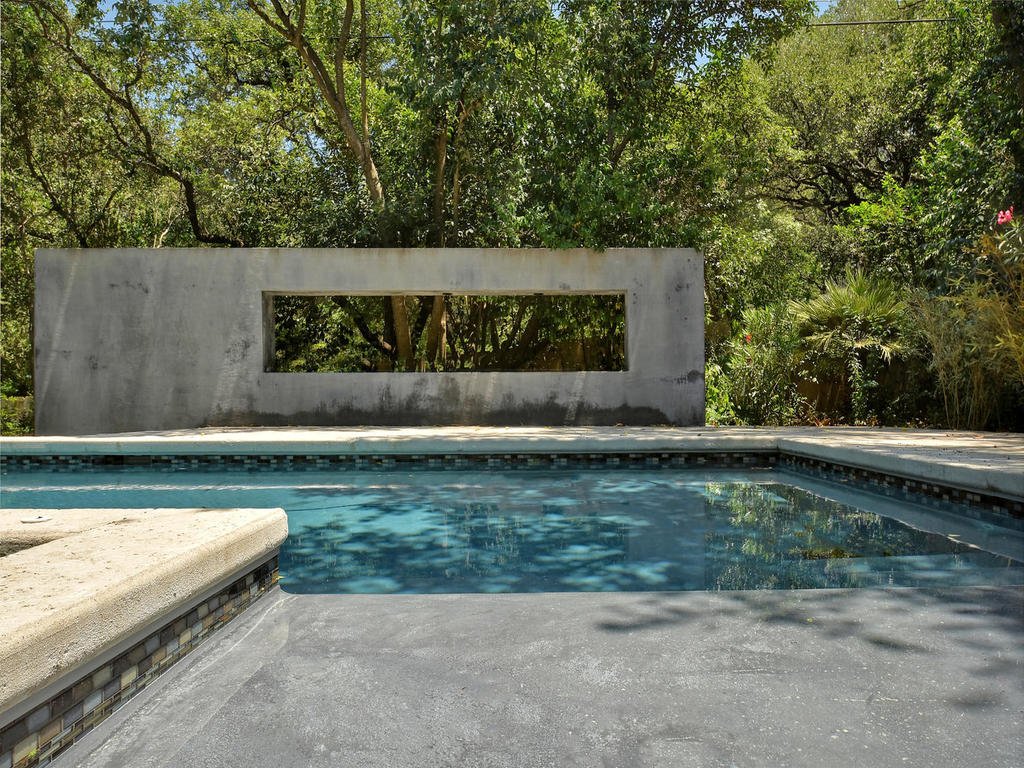 View of a backyard swimming pool with a concrete wall and rectangular window, surrounded by lush green trees and shrubs.