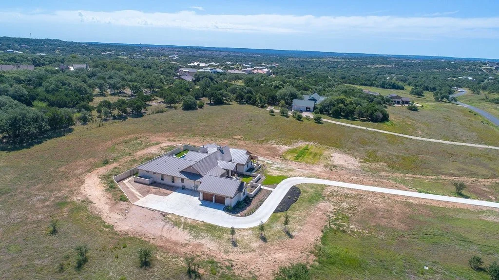A large house under construction on a cleared lot with a driveway, surrounded by open land and trees, in a rural area.