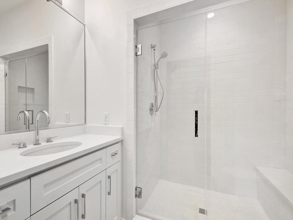 A modern, all-white bathroom with a large mirror, white vanity with dual sinks, and a walk-in shower enclosed by glass. The shower has a handheld showerhead and a tiled bench inside.