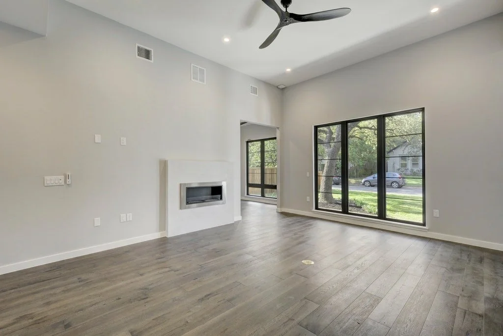 Empty living room with gray walls, hardwood floors, a fireplace, large windows, and a ceiling fan.