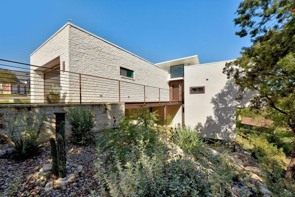 Modern residential house with white brick and stucco exterior, large windows, and a balcony with a metal railing, surrounded by desert plants and rocks under a clear blue sky.