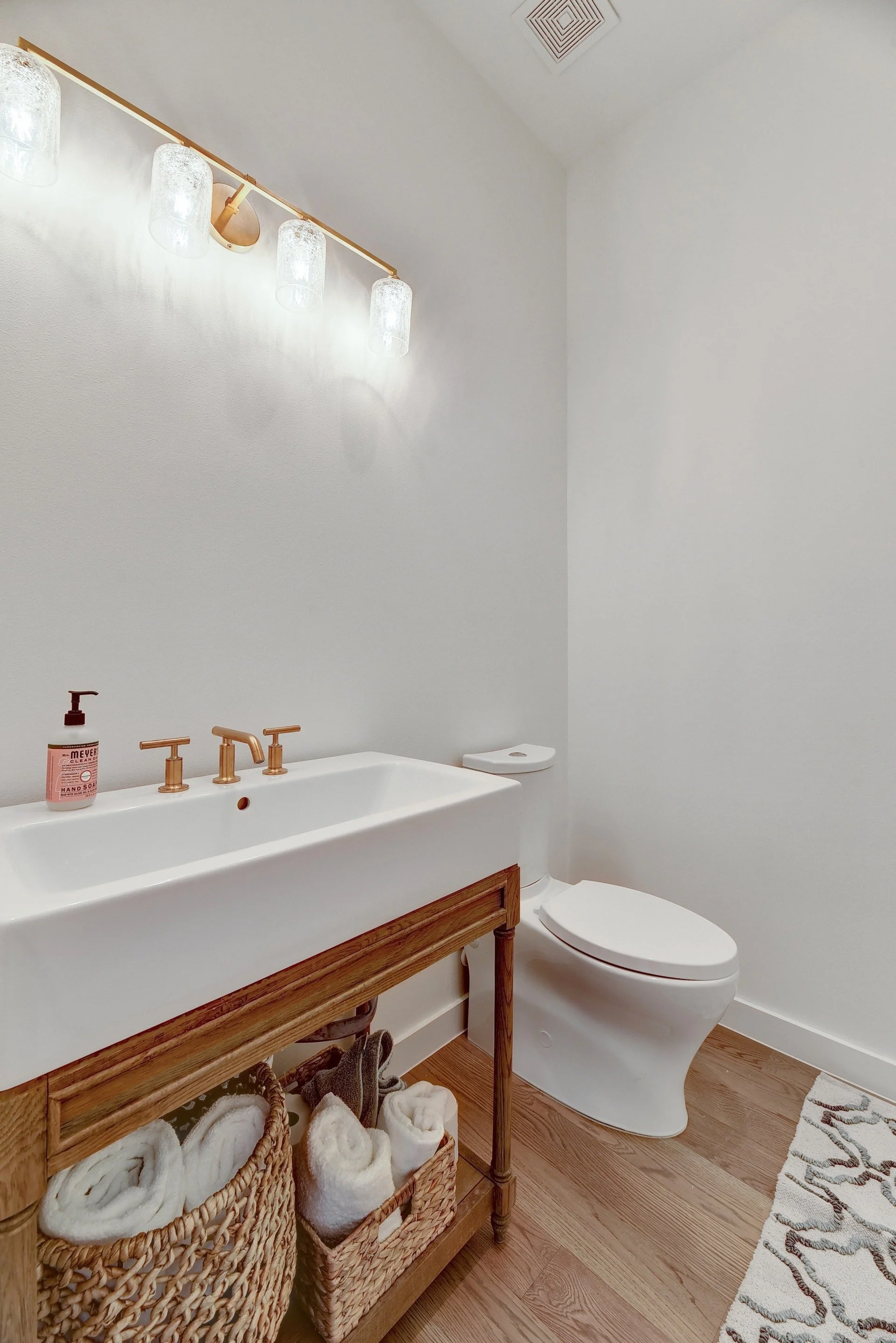 A modern bathroom with a white sink, copper fixtures, a wicker basket with rolled towels, and a white toilet. The walls are painted light gray, and there is a wooden floor with a decorative rug.