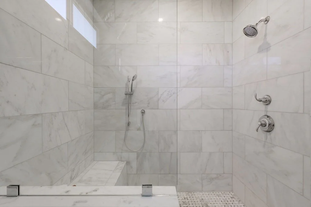 Empty walk-in shower with white marble tiles, a showerhead, a hand-held shower wand, and a small built-in bench.