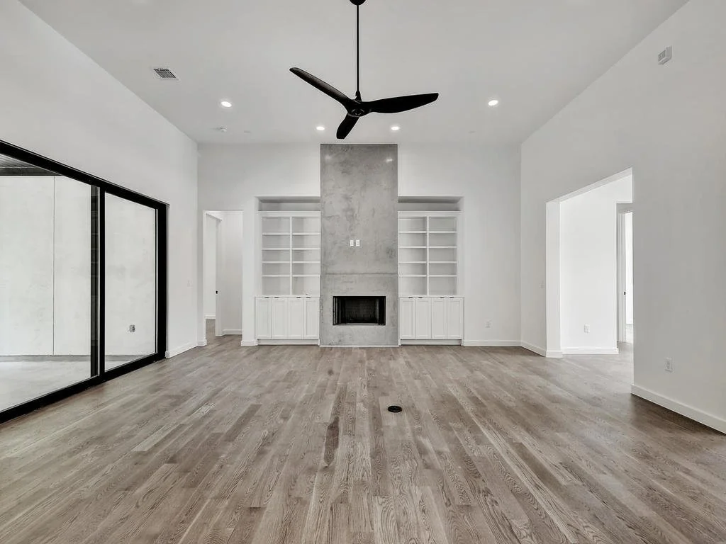 Empty living room with hardwood floors, white walls, built-in shelves around a fireplace, and a ceiling fan.