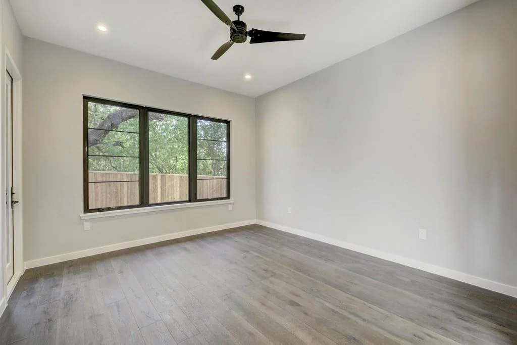 Empty room with large window, ceiling fan, light gray walls, and wooden flooring.