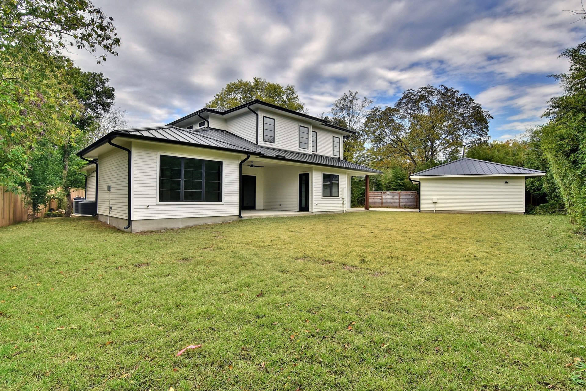 A two-story white house with black trim and a metal roof, situated in a grassy backyard with trees and a partly cloudy sky.