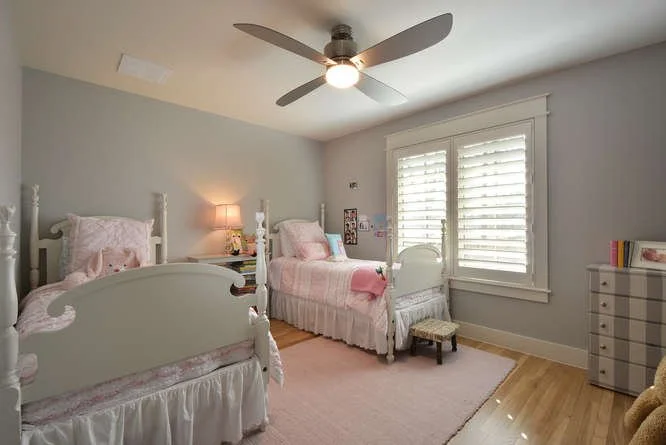 A bright bedroom with two white twin beds, pink bedding, a nightstand with a lamp between the beds, a small white dresser, a window with plantation shutters, and a ceiling fan with a light.