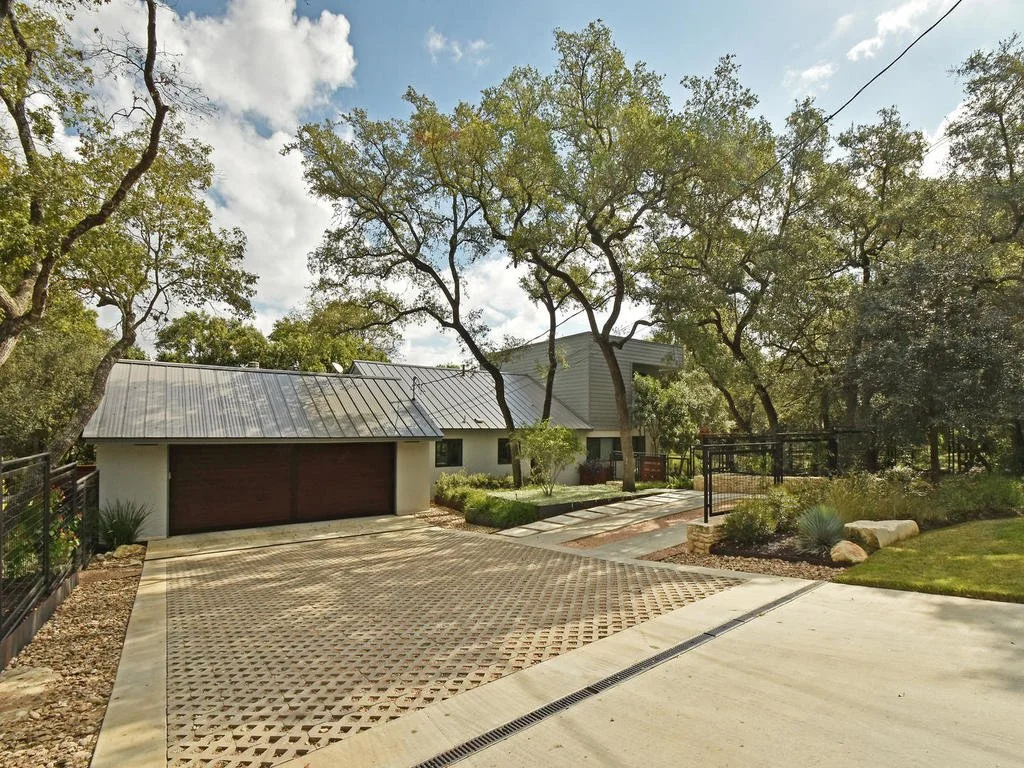 Front yard with trees, a driveway with a patterned surface, a garage with a metal roof, and a house with light-colored siding surrounded by greenery.