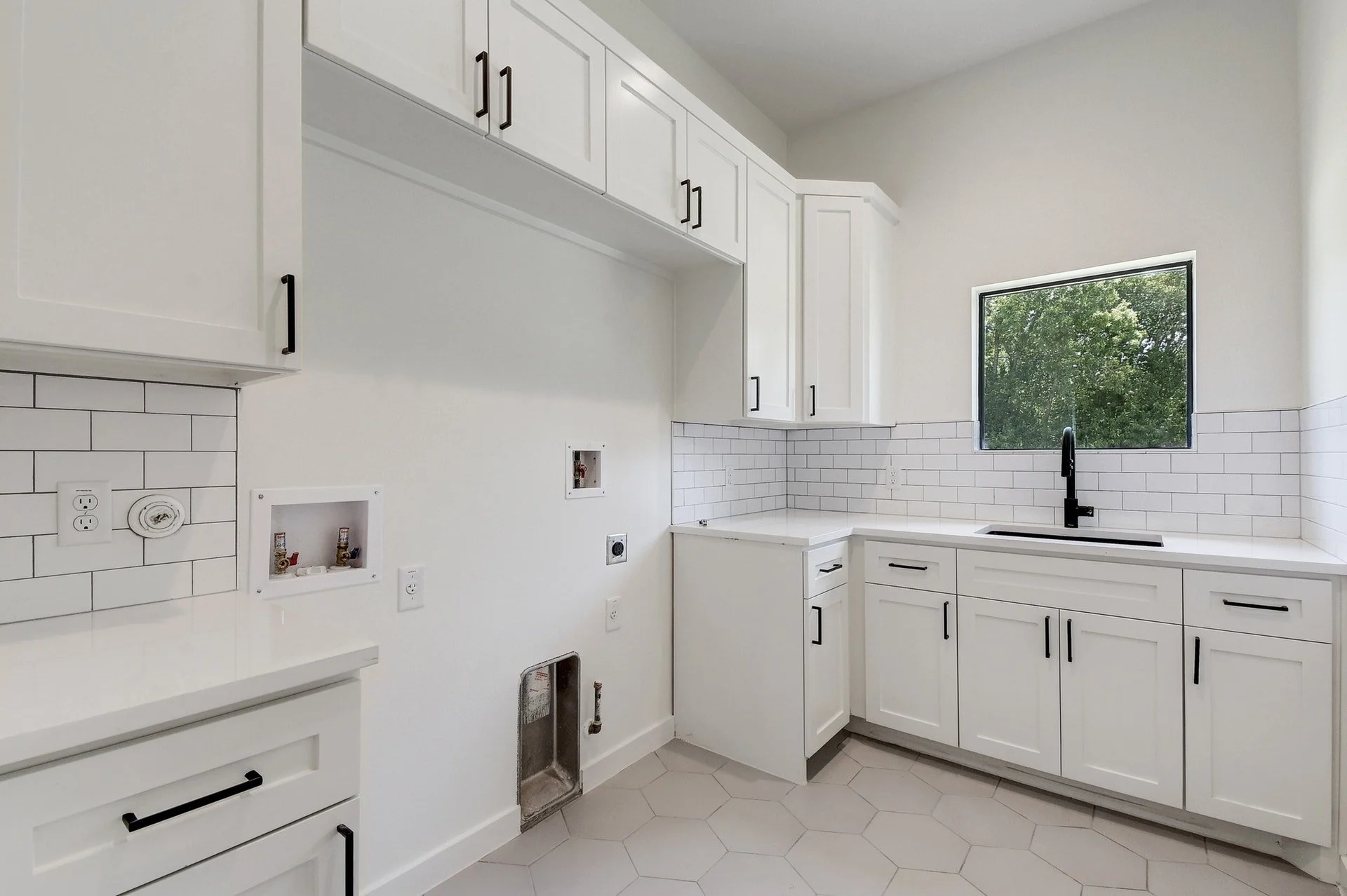 Empty white kitchen with black handles, hexagonal floor tiles, and a window overlooking trees.