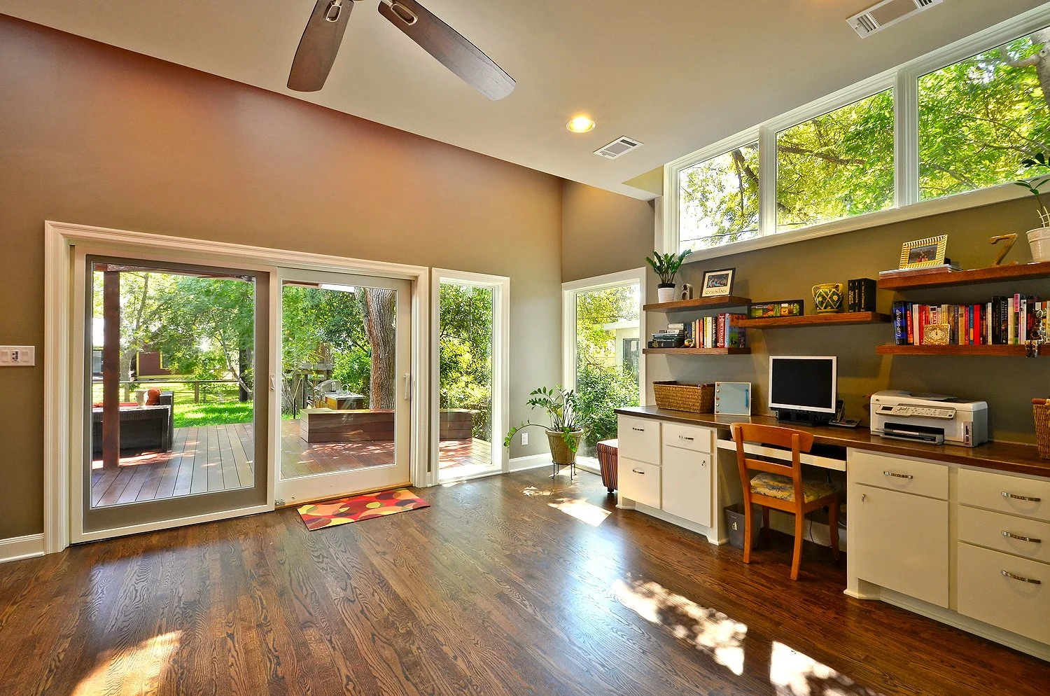 A cozy living room with sliding glass doors leading to a backyard deck. The room has wood flooring, a beige wall, and a green accent wall with wooden shelves filled with books and decor. There's a small desk with a computer and printer, and large win