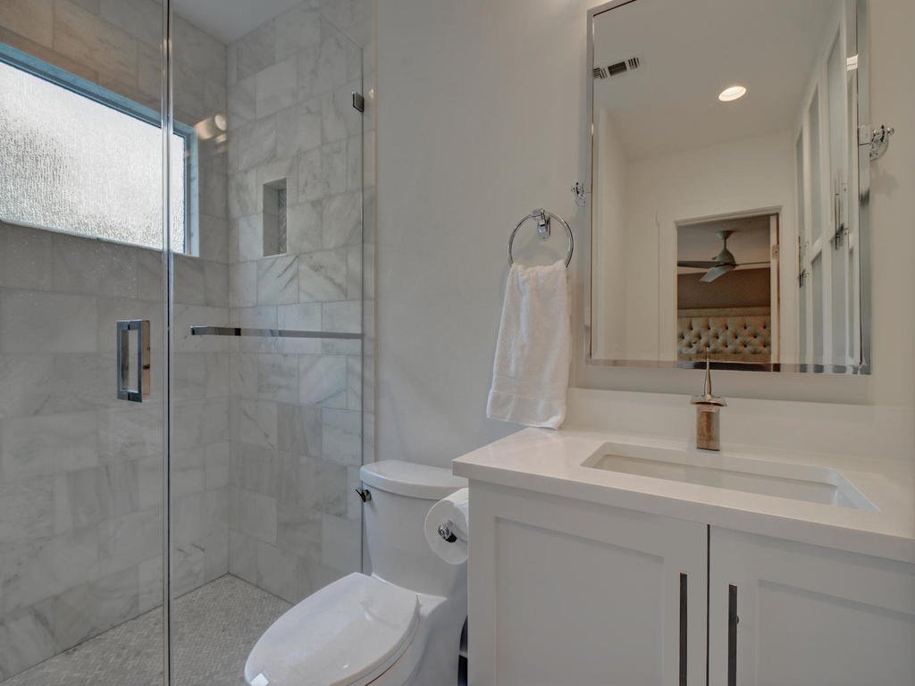 Bathroom with a shower area and a vanity with a mirror, sink, and cabinet. The shower has frosted glass and white marble tiles. There is a window in the shower area, and a towel hangs on a ring next to the mirror.