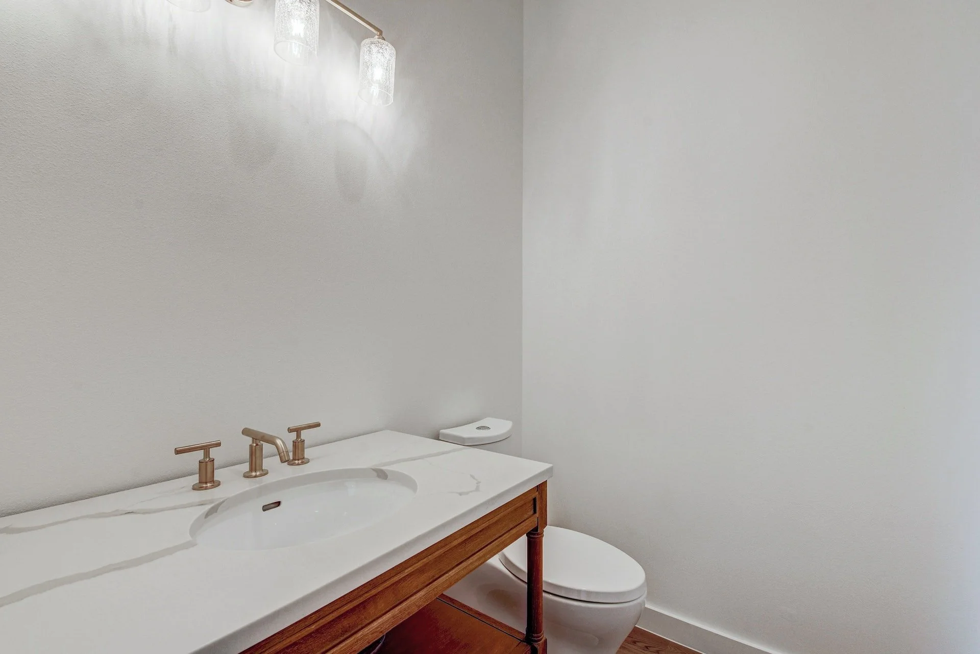 Bathroom with white walls, wooden vanity with a marble countertop, gold fixtures, and a toilet in the background.
